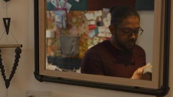 Medium shot of mirror, black man wearing glasses arrives, looks into it, takes his coat, puts coat on and adjusts it whilst looking into mirror, looks down, takes face mask, adjusts it video