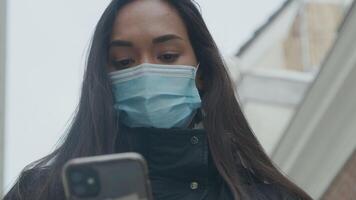 Head shot of young mixed race woman with face mask, standing outside with phone video