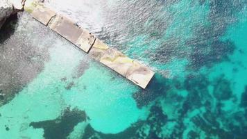 Bird's-eye view of pier extending over Turquoise Mediterranean Sea in Anchor Bay near Popeye Village in Malta - Rotation Aerial shot video