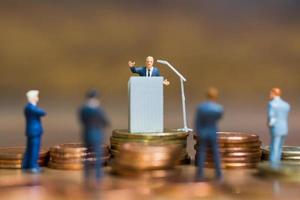 Empresario en miniatura hablando en un podio sobre una pila de monedas, concepto de inversión financiera y empresarial foto