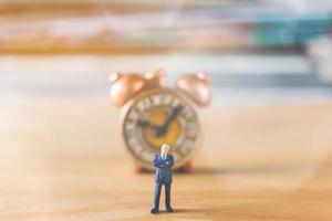 Miniature businessman standing with an old clock on a wooden background photo