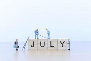 Miniature people working on wooden blocks with the word July on a wooden floor photo