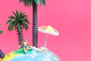 Miniature people wearing swimsuits relaxing on a globe with a pink background, honeymoon concept photo