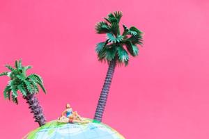 Miniature people wearing swimsuits relaxing on a globe with a pink background, honeymoon concept photo