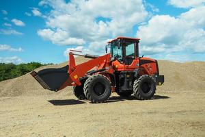 A red-black front-end loader with small wheels against the background of a large pile of stone sand photo