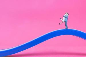 Miniature couple hugging on a bridge with a pink background photo