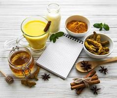 Latte de cúrcuma dorada en un vaso, especias y libro de recetas sobre fondo de madera blanca foto