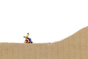 Hombre en miniatura en silla de ruedas sobre un recorte de cartón aislado sobre un fondo blanco. foto