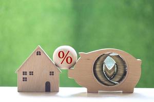 Stack of coins in a wooden piggy bank, a model house, and a percentage symbol on a natural green background photo