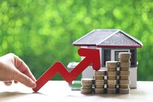 A woman's hand holding a red arrow on a stack of coins money with a model house on a natural green background, business investment and real estate concept photo