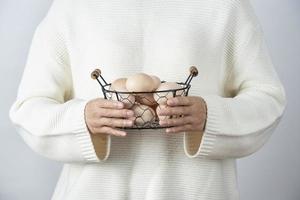 Manos femeninas sosteniendo una canasta metálica llena de huevos de gallina cruda contra una pared gris foto