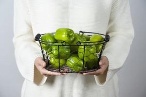 Female hands holding a metallic basket full of green bell peppers against a gray wall photo