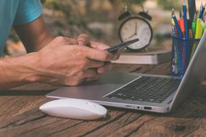 A man is using his phone while sitting at a table with a laptop photo