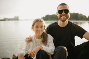 Un padre joven con barba está sentado con su hija en la costa del lago. foto