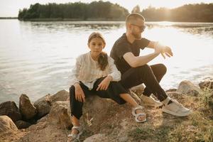 A young father with a beard and sunglasses is sitting on stones with his daughter and checking the smartwatch in the evening photo