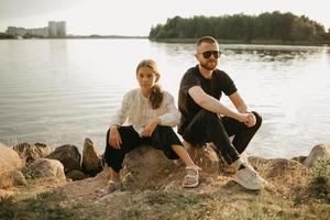 A young father with a beard and sunglasses is sitting on stones with his daughter on the coast photo