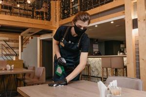 A kind waitress who wears a black medical face mask and disposable medical gloves is holding a bottle with sanitizer and cleaning tables with a rag in a restaurant