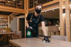 A kind waitress who wears a black medical face mask and disposable medical gloves is holding a bottle with sanitizer and cleaning tables with a rag in a restaurant