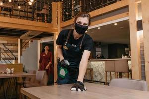 A kind waitress who wears a black medical face mask and disposable medical gloves is holding a bottle with sanitizer and cleaning tables photo