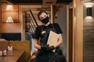 A waitress in black disposable medical gloves wears a medical face mask holding a wooden menu in a restaurant