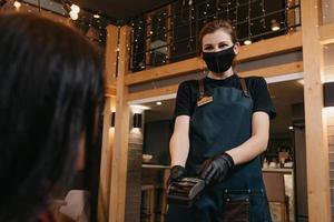 A waitress wears a medical face mask and disposable medical gloves handing a wireless payment terminal to a female client photo
