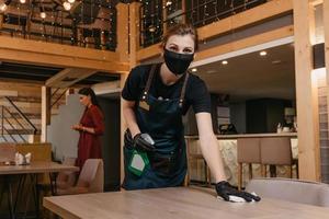 A kind waitress wears a black medical face mask and disposable medical gloves cleaning tables photo