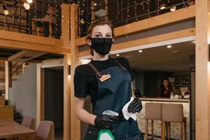 A kind waitress wears a black medical face mask and disposable medical gloves holding a bottle with sanitizer and cleaning tables