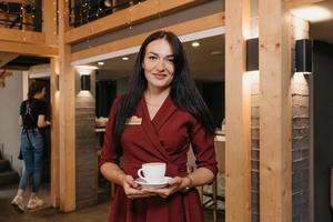 A female restaurant manager wears a black face mask and disposable gloves holding a cup of coffee in a restaurant photo