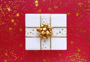 Caja de regalo blanca con lazo dorado y estrellas sobre un fondo rojo. foto