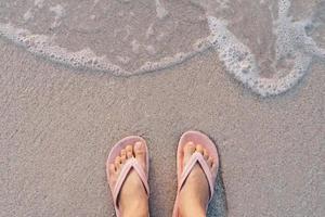 Top view of a woman's feet wearing slippers standing on a sandy beach with sea waves photo