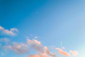 Nature background of blue sky with pink clouds, airplane trail, and early moon