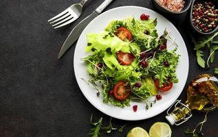Fresh green mixed salad bowl with tomatoes and microgreens on black concrete background photo