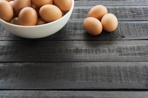 Huevos de gallina en un recipiente blanco y tres huevos de gallina afuera sobre una mesa de madera negra foto