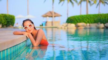 Mujer asiática sonríe mientras toma el sol en la piscina con palmera frente al mar video