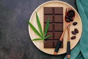 Hoja de cannabis con chocolate negro, hojas de plantas y utensilios de madera sobre un fondo de hormigón oscuro foto