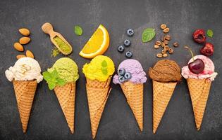 varios sabores de helado en conos de arándano, té verde, pistacho, almendra, naranja y cereza sobre un fondo de piedra oscura. concepto de menú de verano y dulces foto