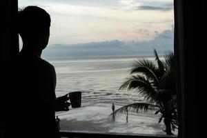 Silhouette of man holding the coffee cup and looking at the sea from the bedroom photo