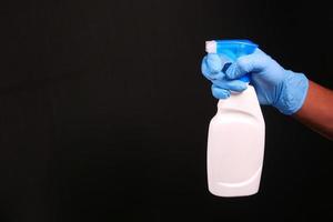 Mano en guantes de goma azul sosteniendo una botella de spray aislado en negro foto