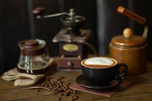 Closeup top view of hot cappucino cup with selective focus on coffee beans on wooden table with blurred grinder background photo