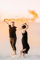 Guy and a girl in black clothes hug and run on the white sand with orange smoke photo