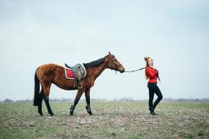 Red-haired jockey girl in a red cardigan and black high boots with a horse photo