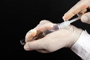 El médico recoge el medicamento de una ampolla en una jeringa con guantes estériles blancos sobre un fondo oscuro foto
