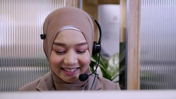 Beautiful woman in hijab at the computer with headset, distant work, call center. Confident islam woman worker in call center crossed arms face camera smiling joyful operator and helps with the order. video