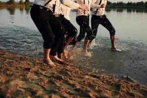 Men's feet in the water. Men in costumes run on water. They are having fun, playing, and splashing water around them. Summer. Group of happy young men's feet splash water in the sea and spraying at the beach photo