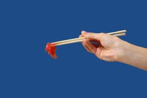 Close up of woman's hand holding ginger for sushi with wooden chopsticks isolated on blue background. Copy space. photo