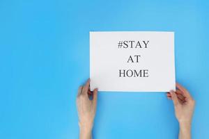 Stay at home concept. Woman hand holding a paper with text, hashtag stay at home, isolated on blue background. Coronavirus, COVID-19, self-quarantine, isolation. photo