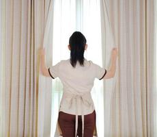 Joven sirvienta abriendo cortinas en la habitación del hotel