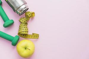 Dumbbells, water bottle, measuring tape and an apple on pink background photo