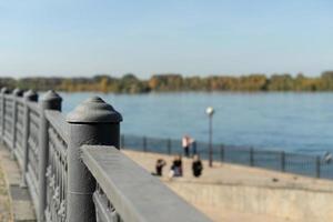 Urban landscape of a metal fence next to a body of water with blurred people in the background and a clear blue sky in Irkutsk, Russia photo
