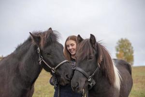 Smiling young woman standing in a field with her two Icelandic horses